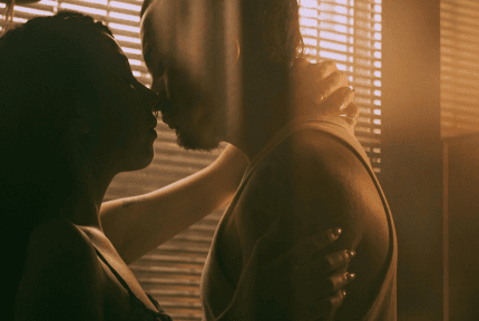 Silhouettes of two people leaning in as if to kiss, sunlit blinds creating a warm atmosphere behind them.