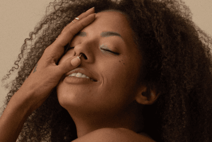 Close-up of a young woman with brown skin, eyes closed, smiling with her head tipped back and hand gently touching her face.
