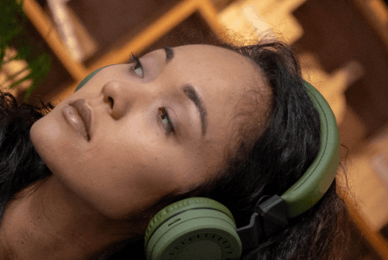 Young woman with brown skin, gazing off thoughtfully while wearing dark green headphones, with cozy mood lighting in the background.