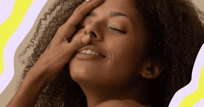 Close-up of a young woman with brown skin, eyes closed, smiling with her head tipped back and hand gently touching her face.